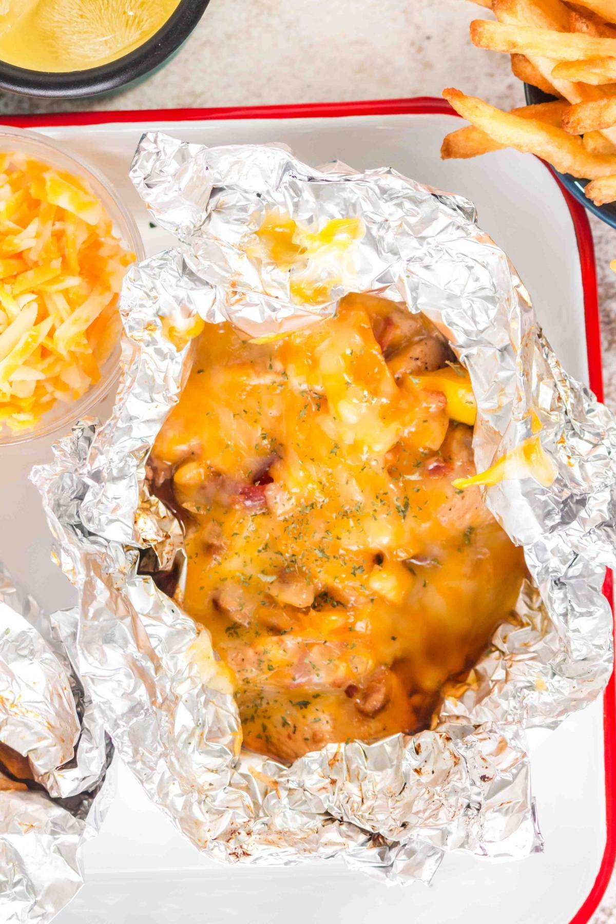 Baked cheesy chicken and bacon in foil, served with shredded cheese and fries on a white tray.