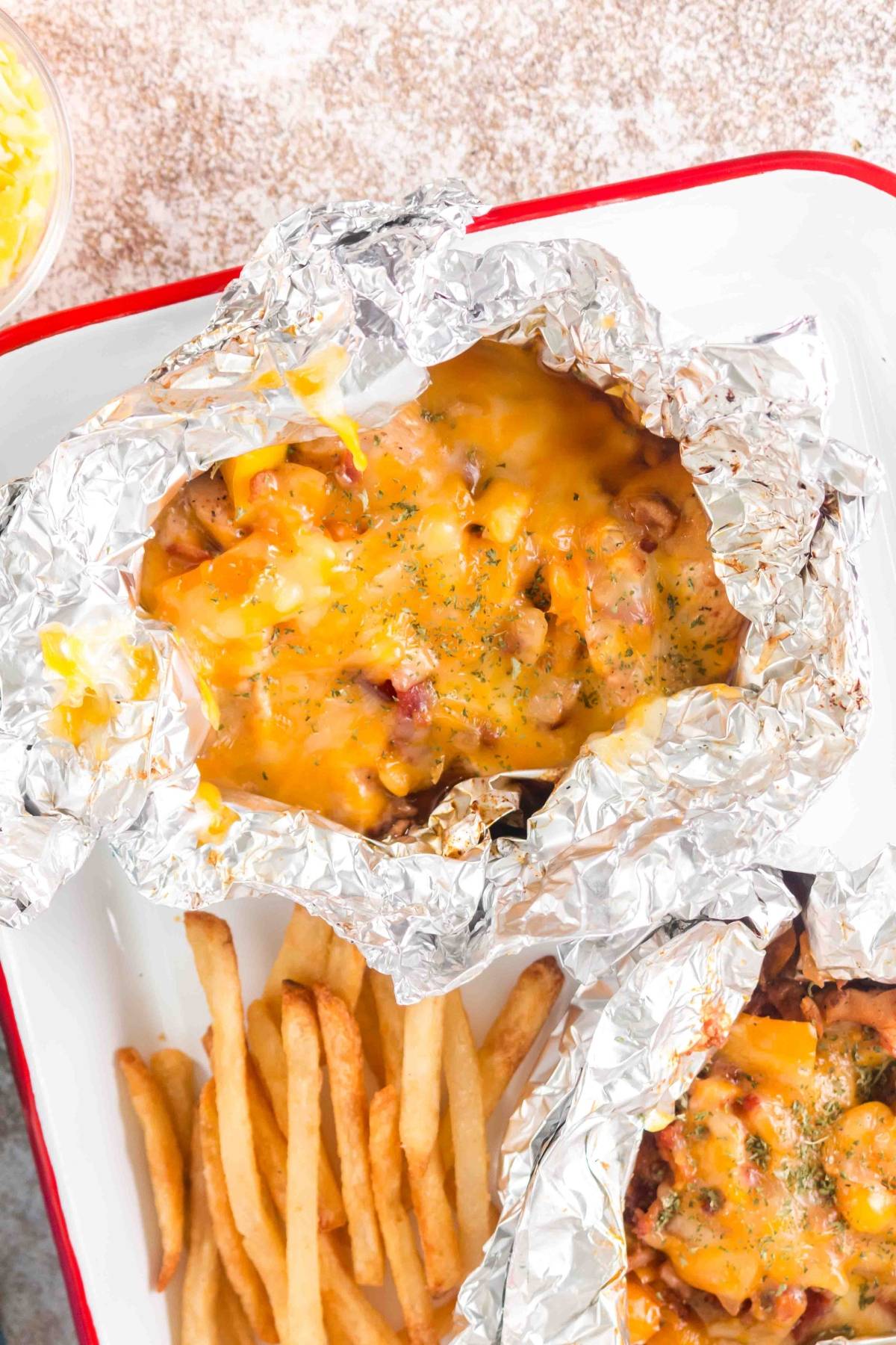 Foil packet with melted cheese and meat next to a serving of French fries on a white tray.