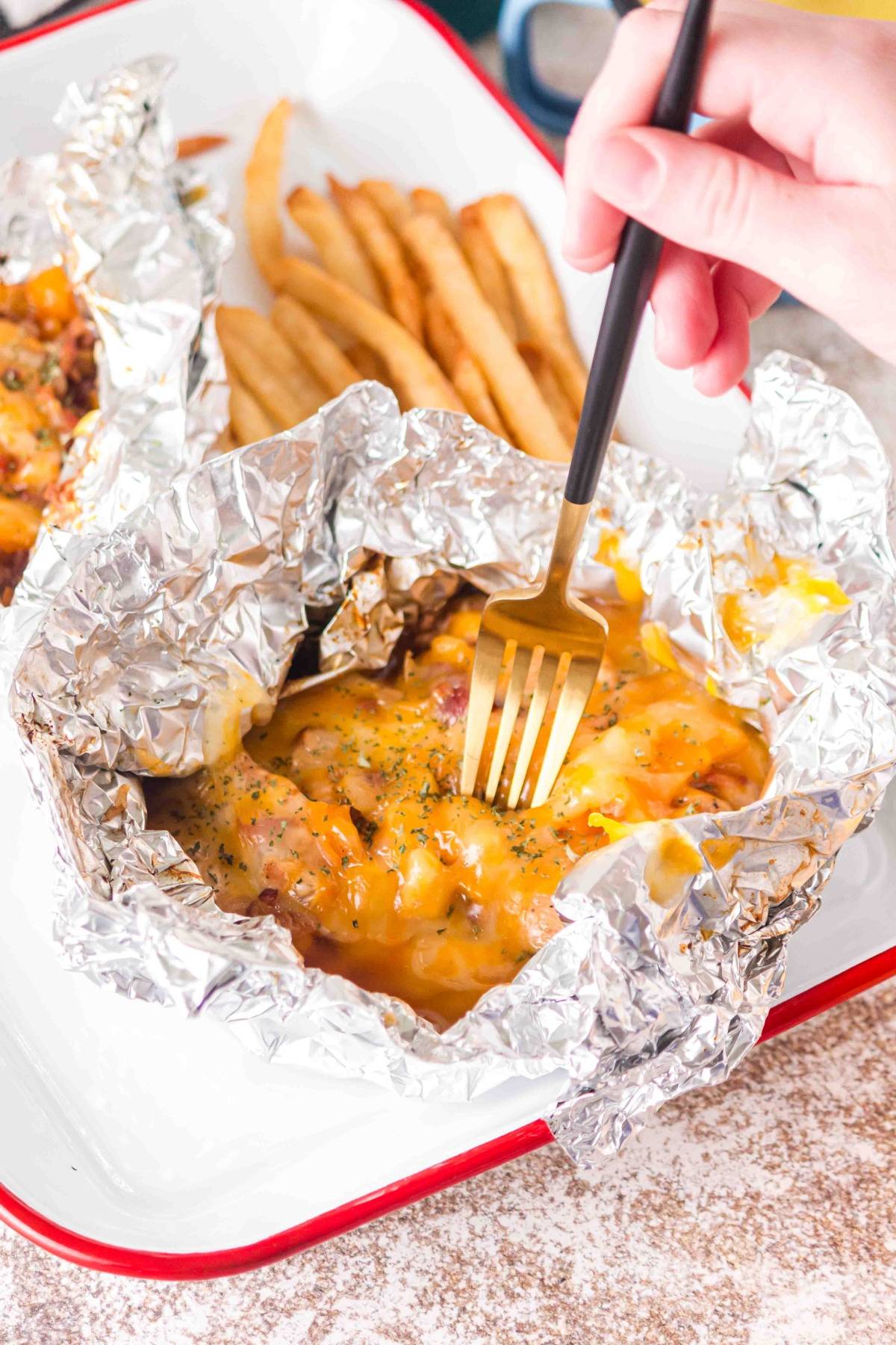 A hand uses a fork to eat cheesy foil packet food, with French fries on the side.