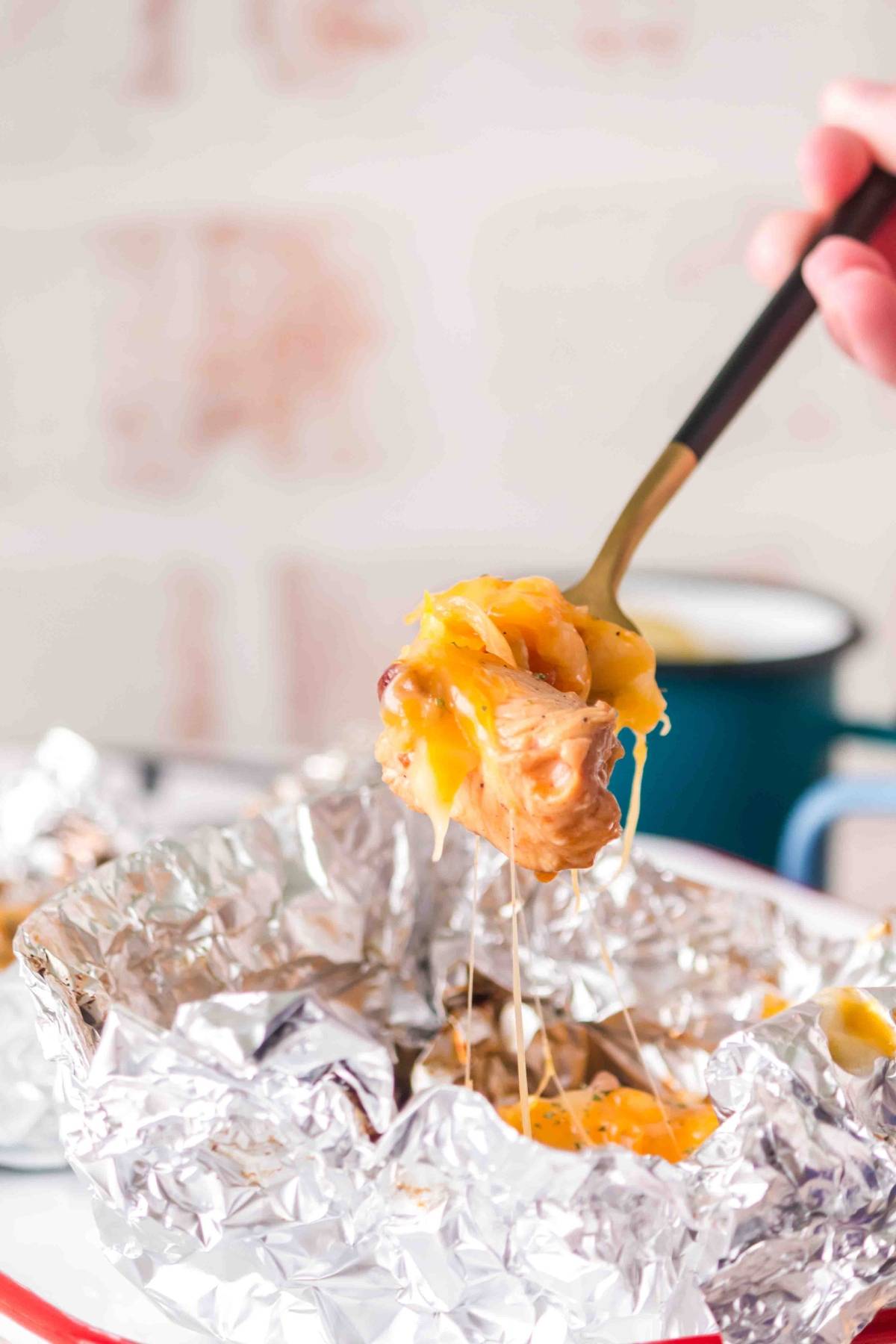 A fork lifts cheesy chicken from a foil packet, with melted cheese stretching from the food.