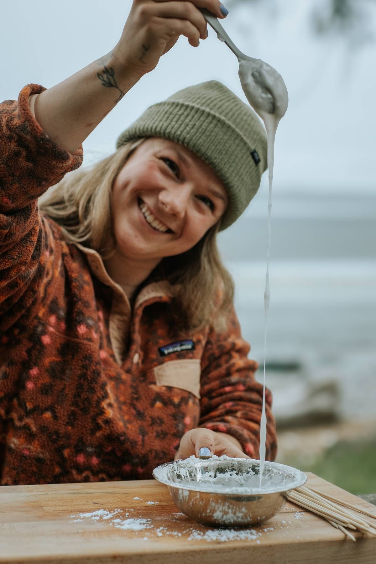 Smiling person in a beanie stretches gooey mixture from a bowl with a spoon at an outdoor table.