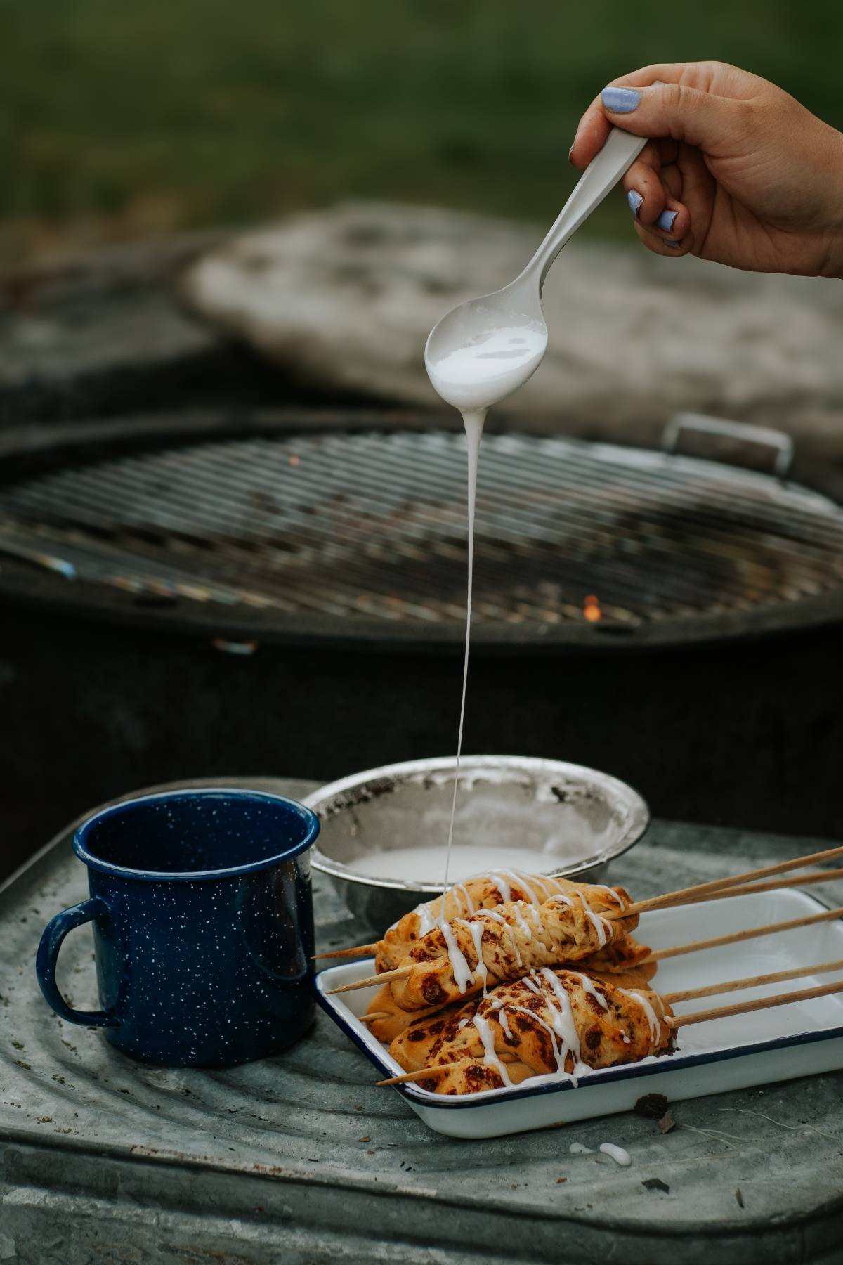 A hand drizzles white icing over skewered pastries next to a blue mug and a grill in the background.
