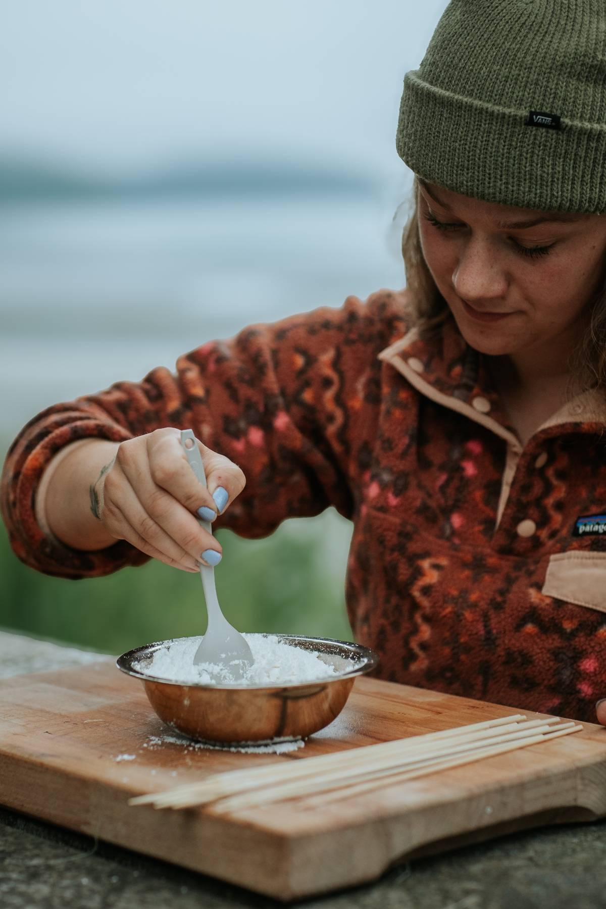 Person in a green beanie stirs white powder in a bowl on a wooden board outdoors.