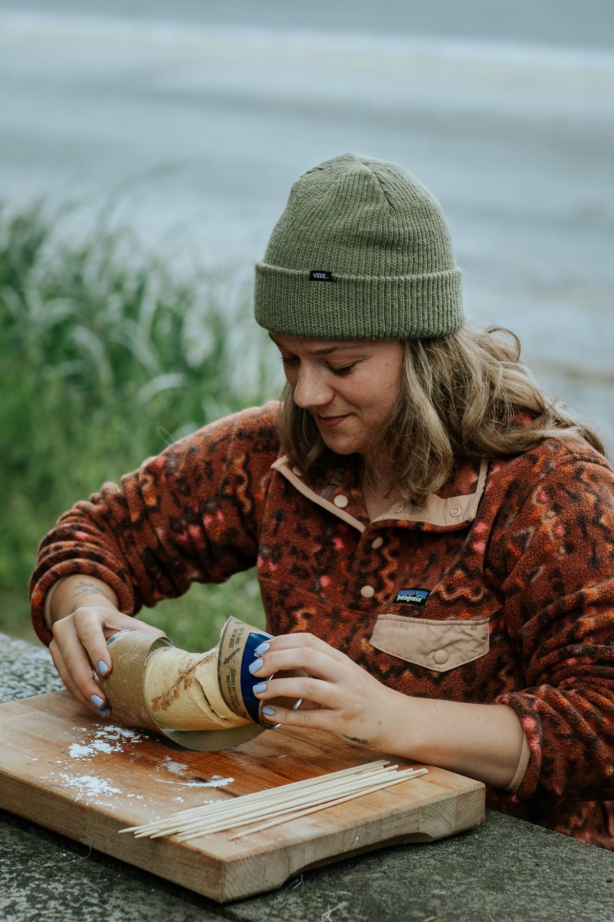 A woman in a beanie opens a can on a cutting board outdoors, with skewers and flour beside her.
