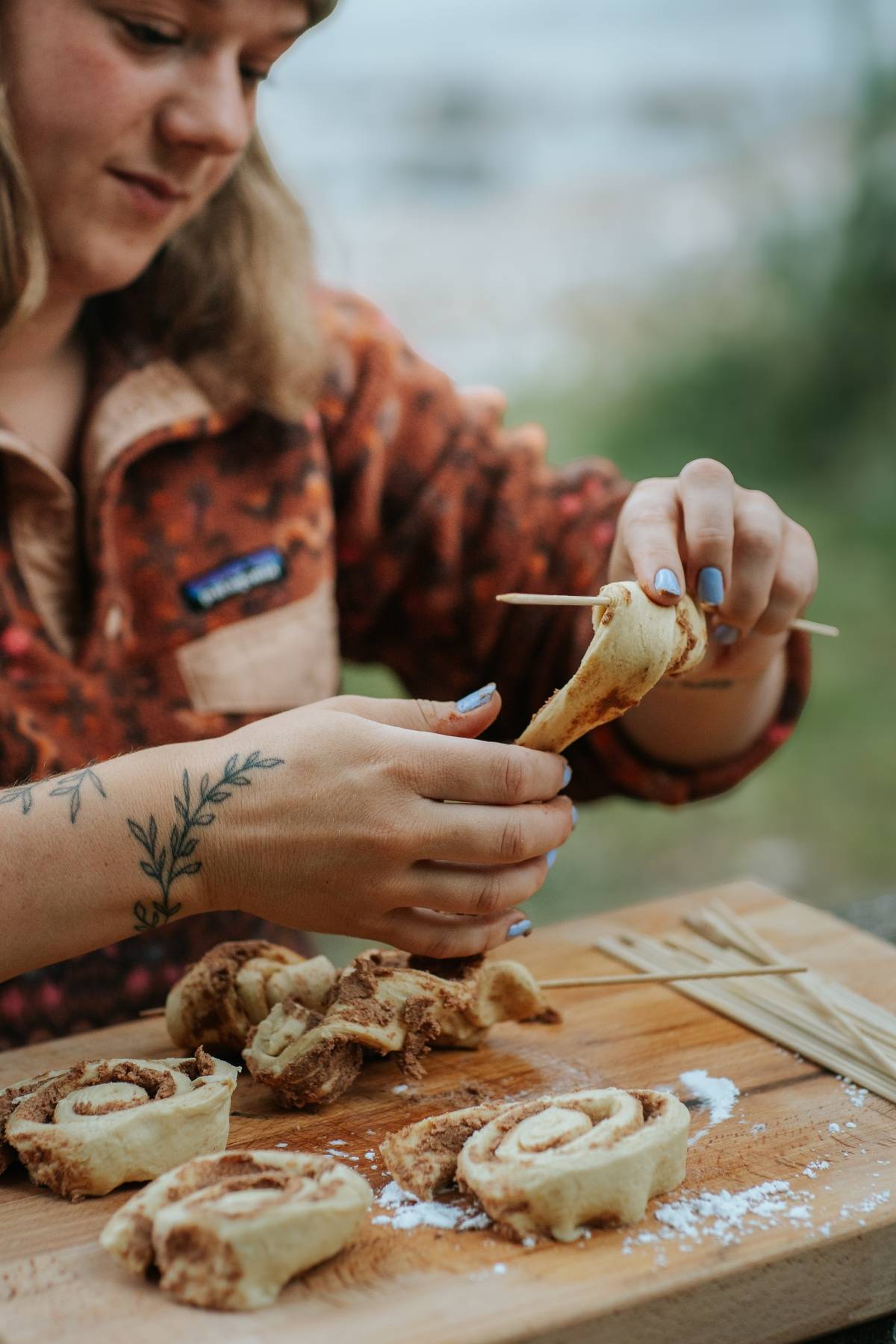 A person wraps cinnamon roll dough around a stick, with more dough rolls and flour on a wooden board.