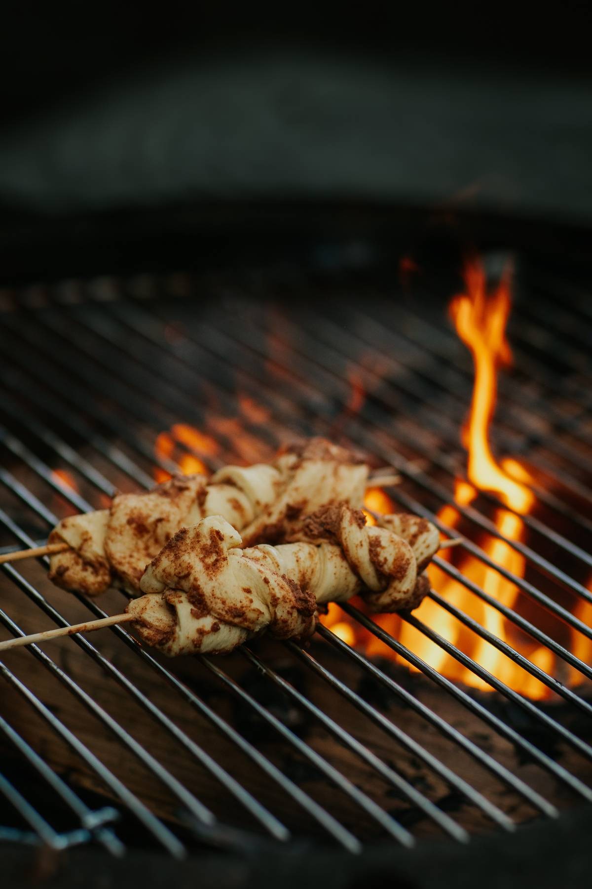 Bread dough twisted on skewers being grilled over an open flame on a barbecue grill.