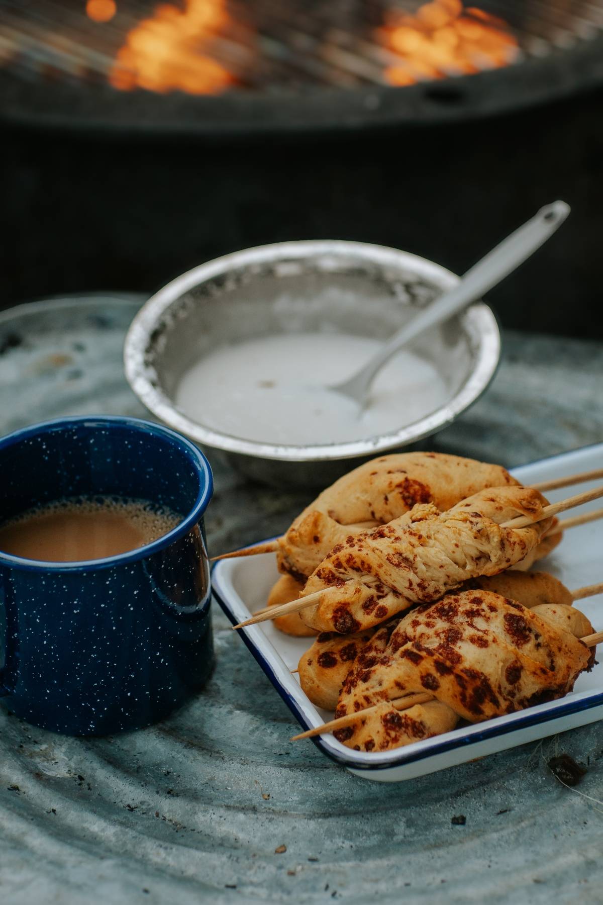 Grilled skewered bread, a blue mug of tea, and a bowl of sauce on a metal surface by a campfire.