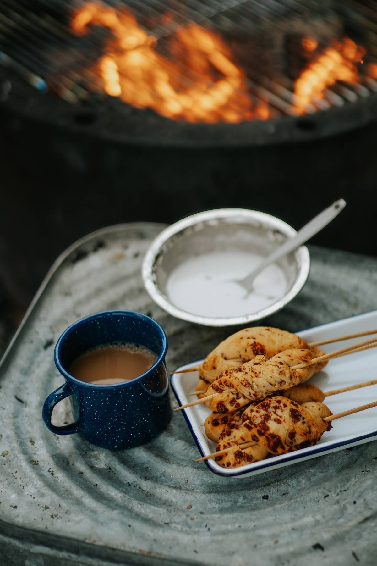 Blue mug of coffee, grilled skewers, and a dish with a spoon on a metal surface by a campfire.