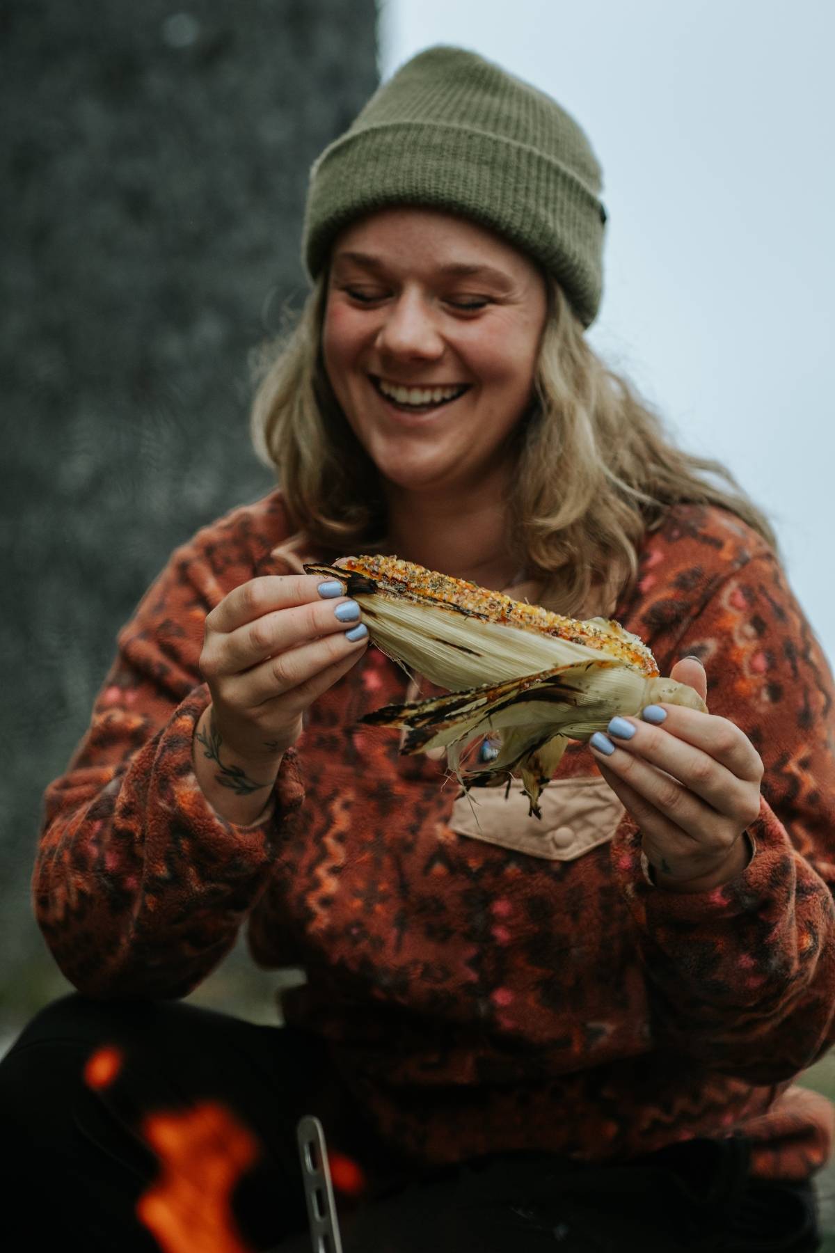 Smiling person in a beanie holds a grilled ear of corn outdoors, wearing a patterned sweater.