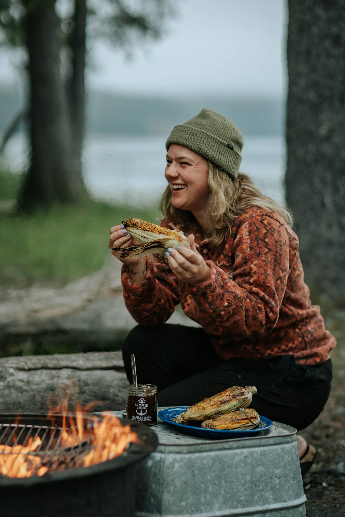 A smiling person in a beanie eats grilled corn by a campfire near a lake, holding two ears of corn.