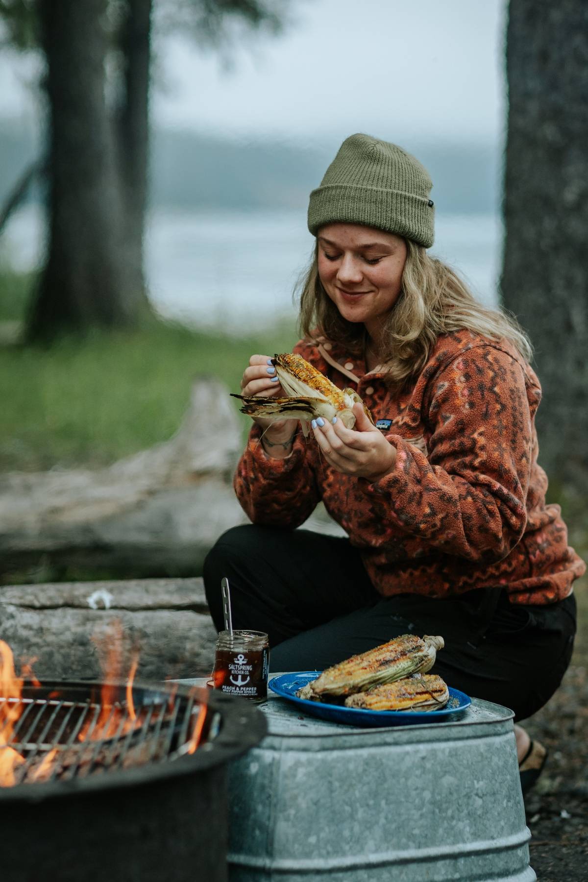 A woman in a beanie eats grilled corn by a campfire in a forested outdoor setting.