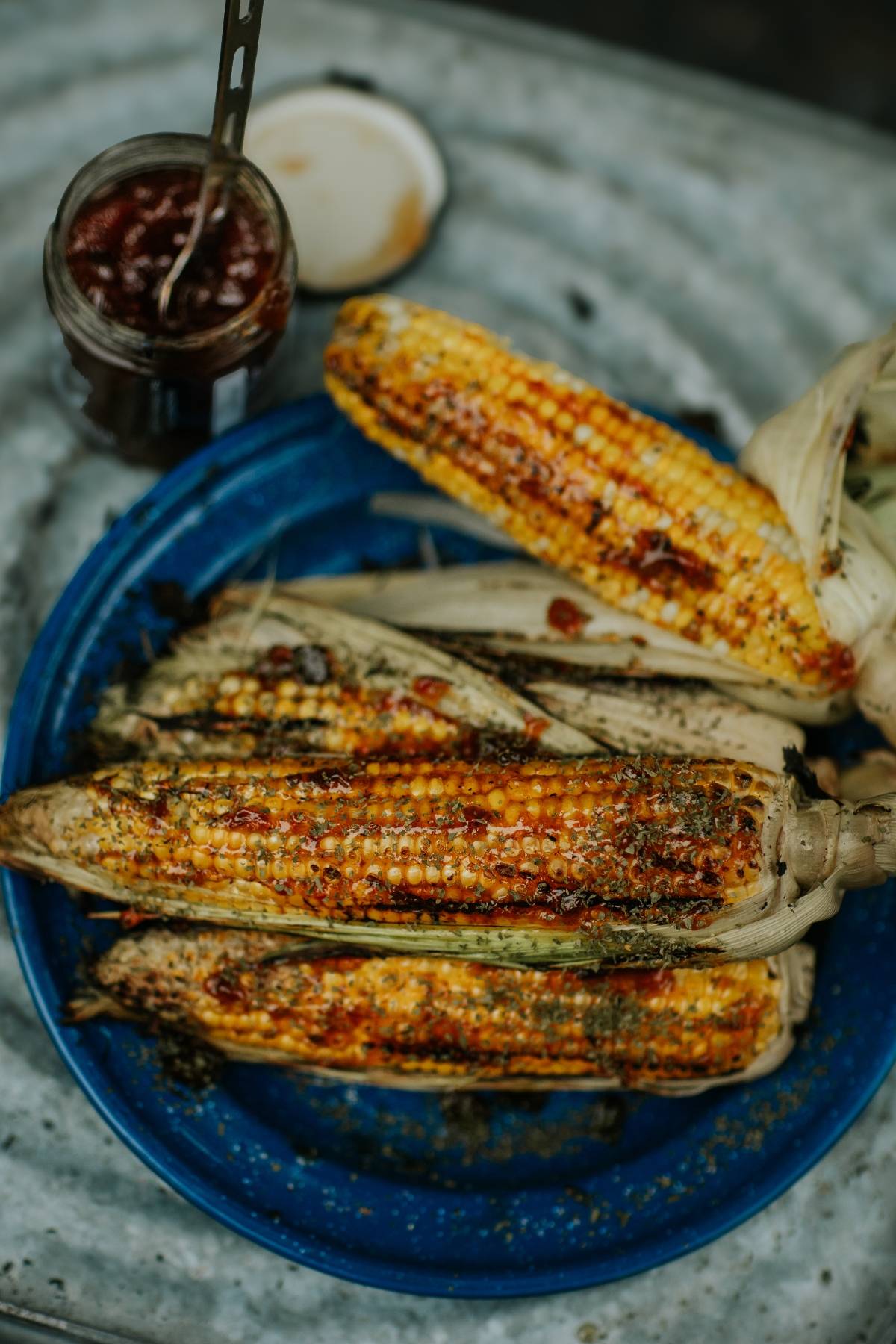 Grilled corn on the cob with spices on a blue plate next to a jar of sauce.