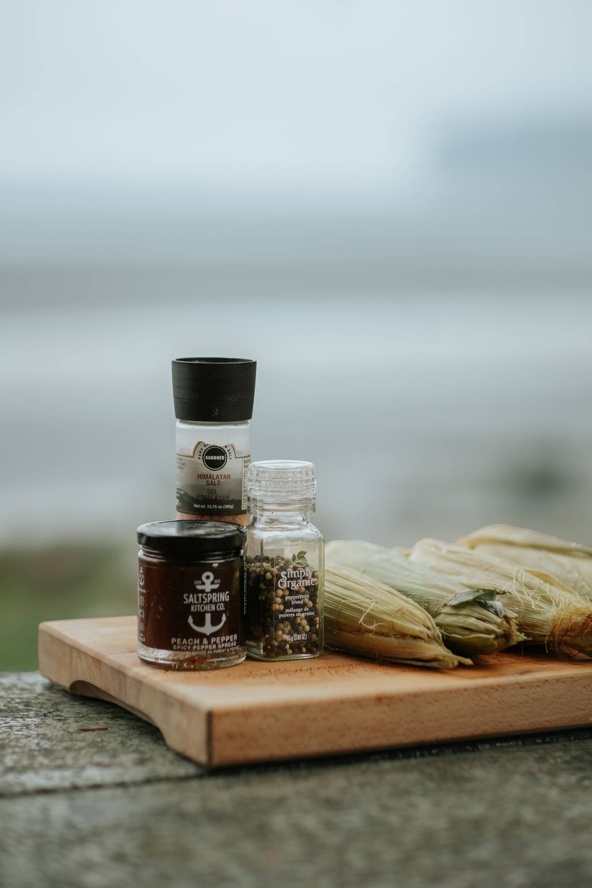 Seasonings and tied corn husks displayed on a wooden board outdoors with a blurred background.
