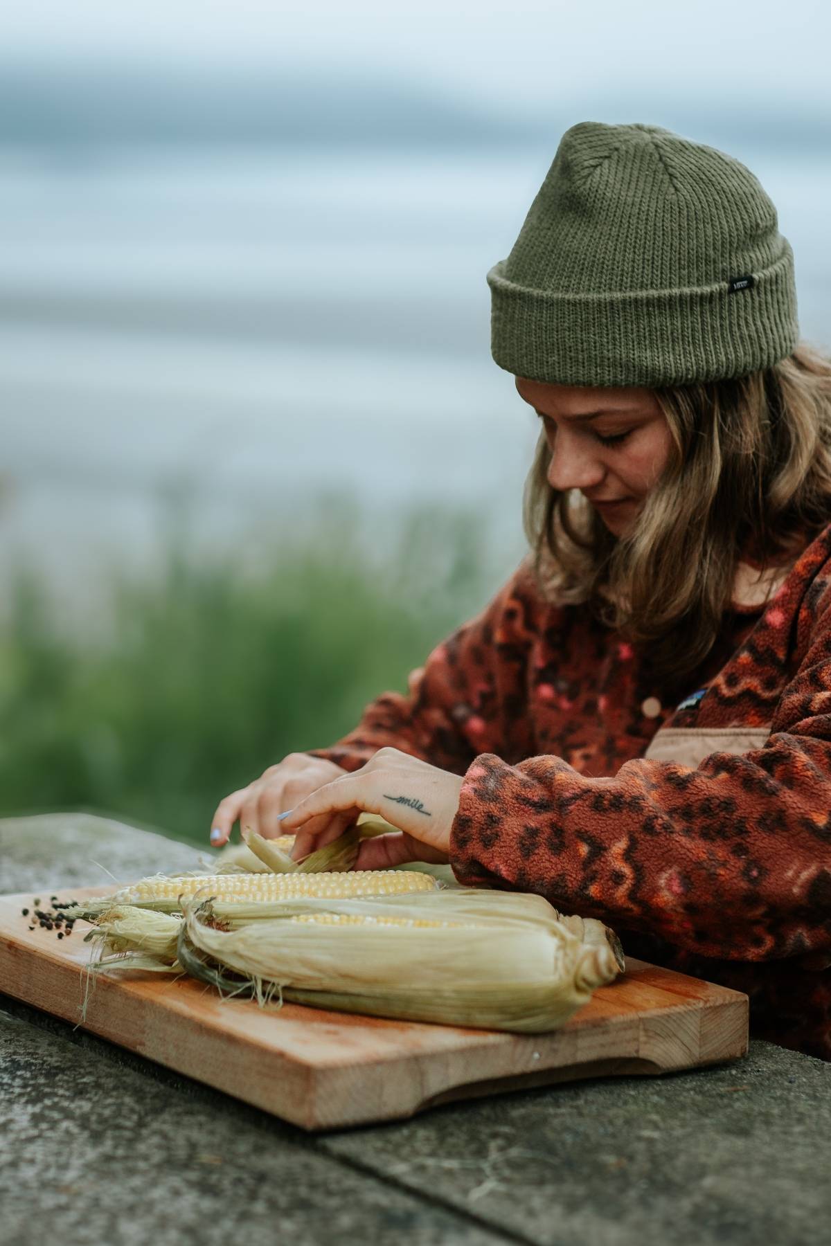 A person in a beanie peels corn on a wooden board outdoors near water.