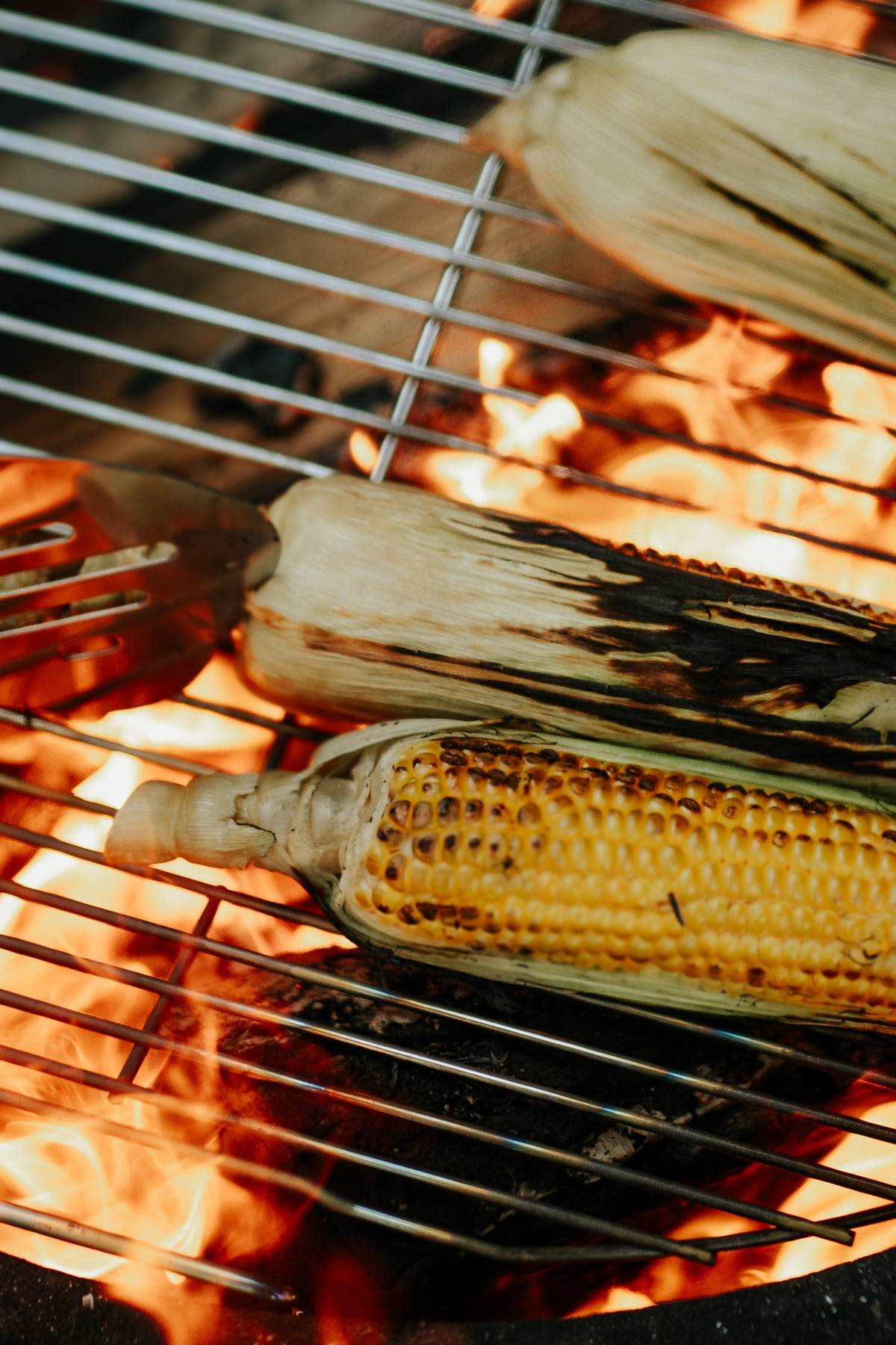 Corn on the cob being grilled over an open flame, with some husks charred on the grill.