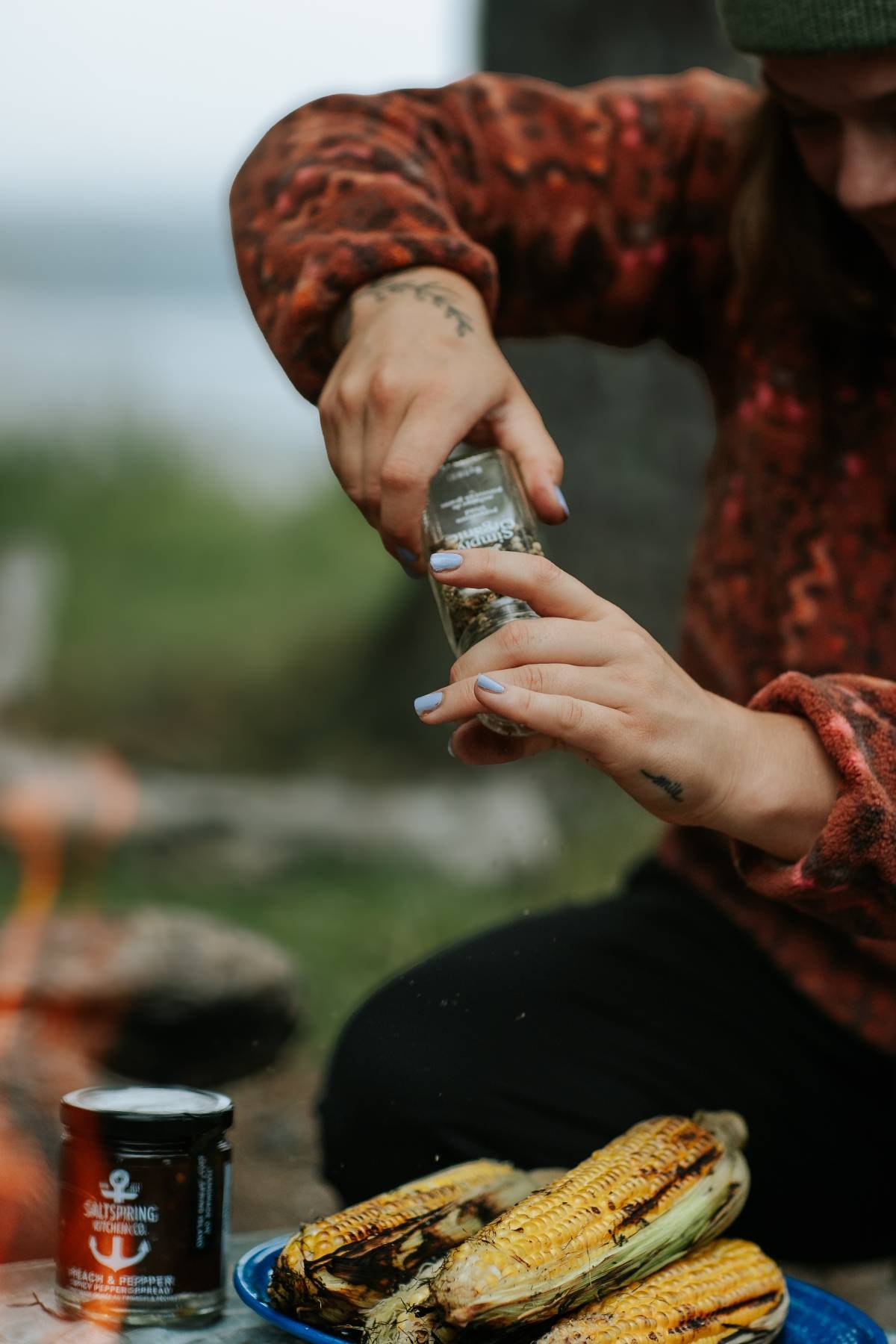 Person seasoning grilled corn on the cob with spices outdoors, with a can of seasoning nearby.