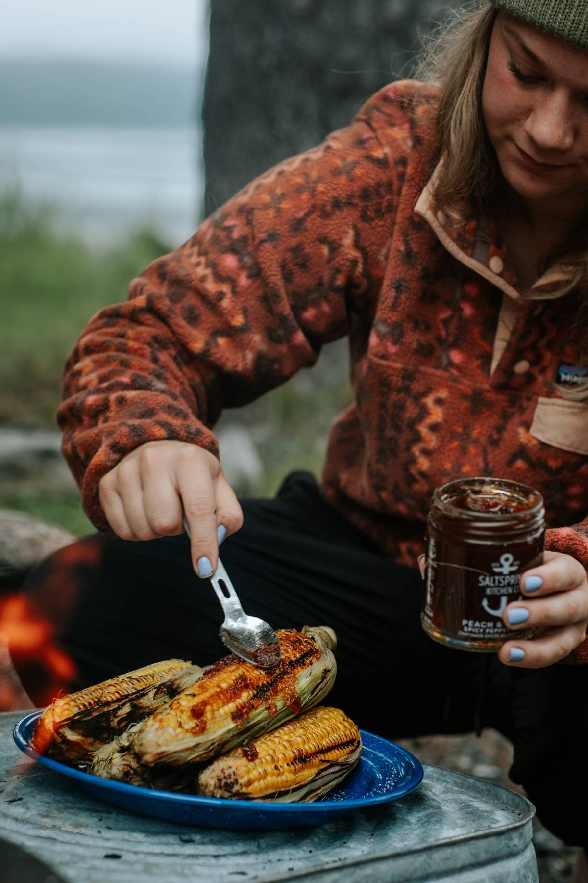 A person spreads seasoning on grilled corn with a spoon next to a campfire outdoors.