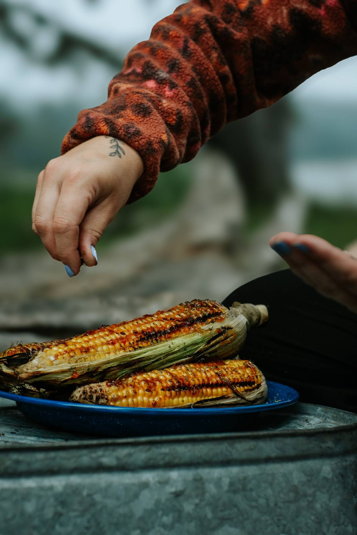 A hand sprinkles seasoning on grilled corn cobs placed on a blue plate outdoors.