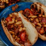 Two grilled hot dog buns filled with campfire hot dogs and beans, tomatoes, and onions on a blue plate.