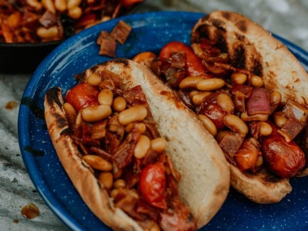 Two grilled hot dog buns filled with campfire hot dogs and beans, tomatoes, and onions on a blue plate.