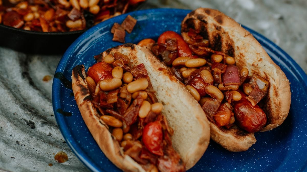 Two grilled hot dog buns filled with campfire hot dogs and beans, tomatoes, and onions on a blue plate.