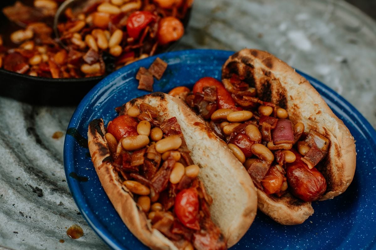 Two grilled buns on a blue plate filled with beans, tomatoes, and vegetables—like a twist on campfire hot dogs and beans—with extra filling warm in the pan behind.