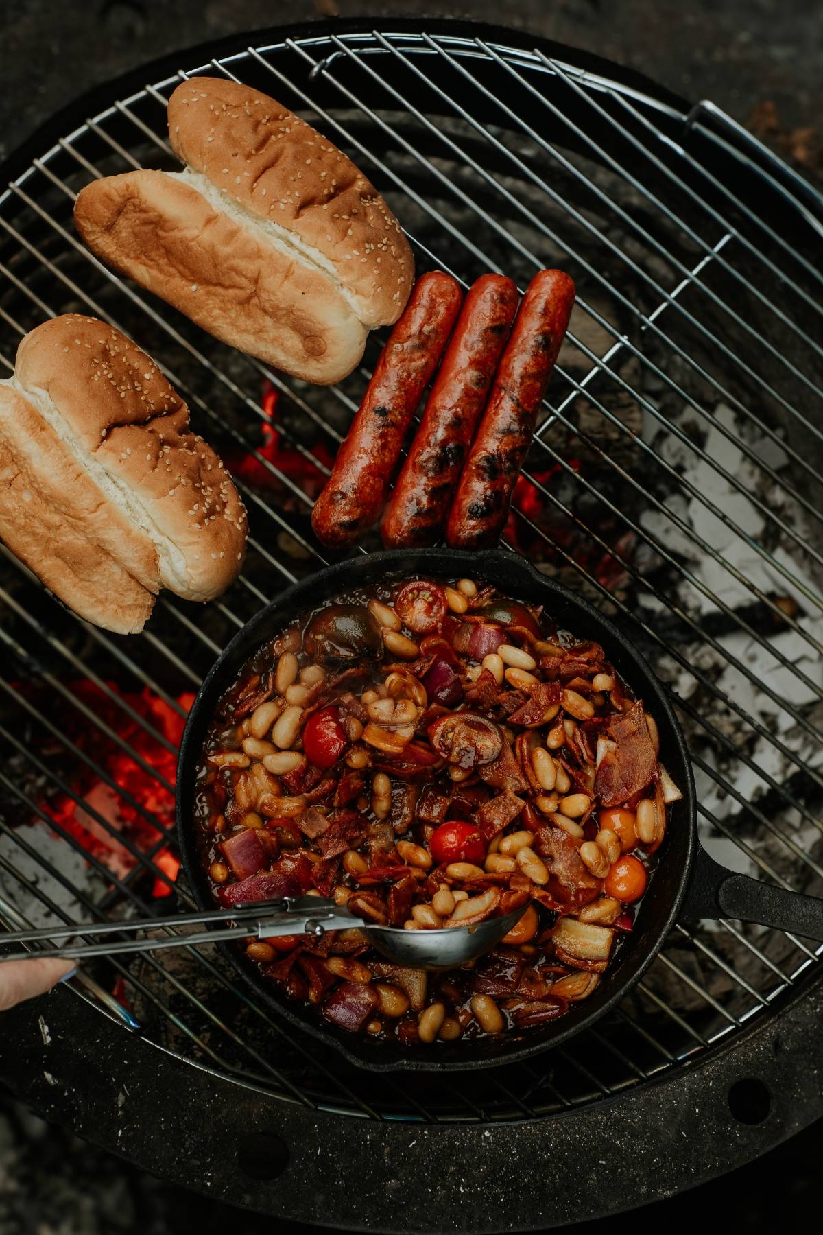 Hot dogs, buns, and a skillet of bean stew cooking over an outdoor grill with glowing coals underneath.