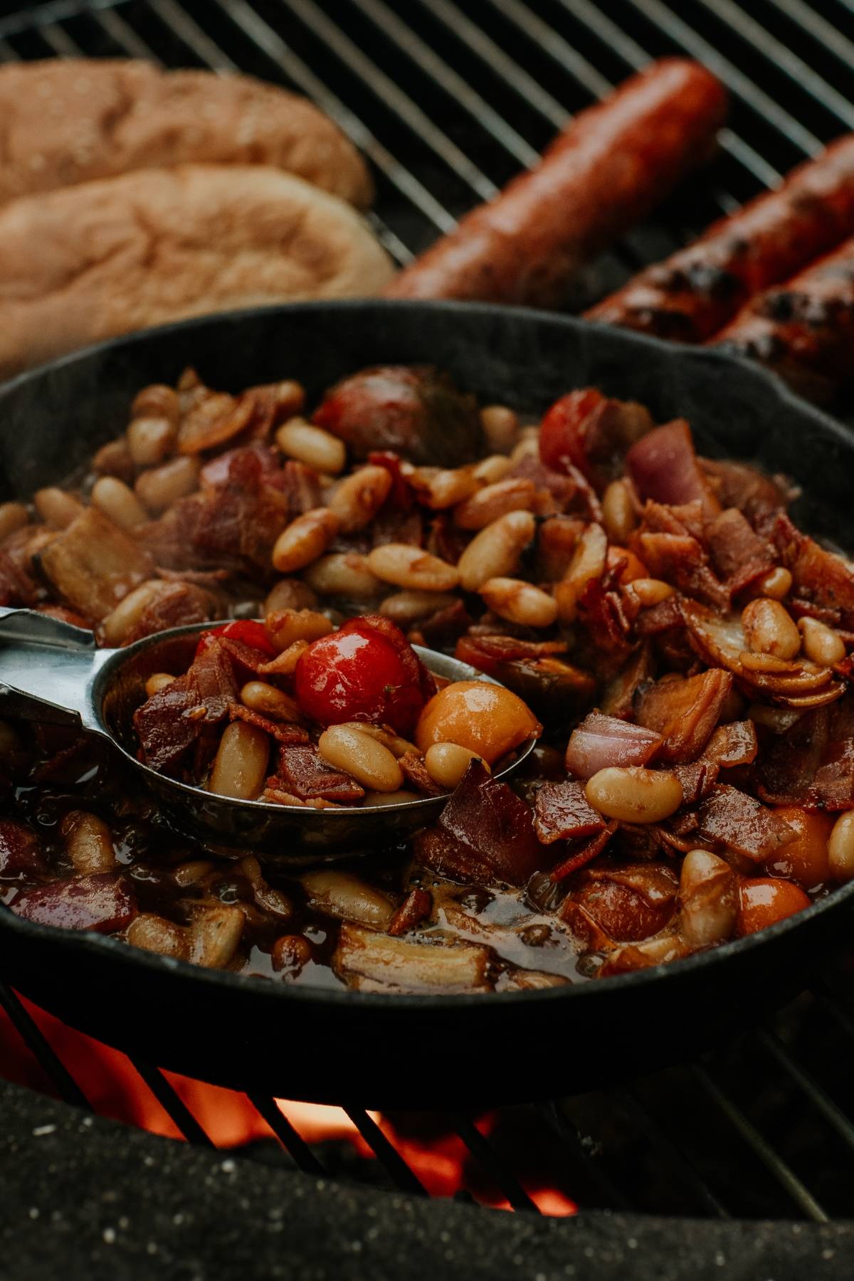 Skillet of baked beans with bacon and tomatoes cooking on a grill, with buns and sausages in the background.