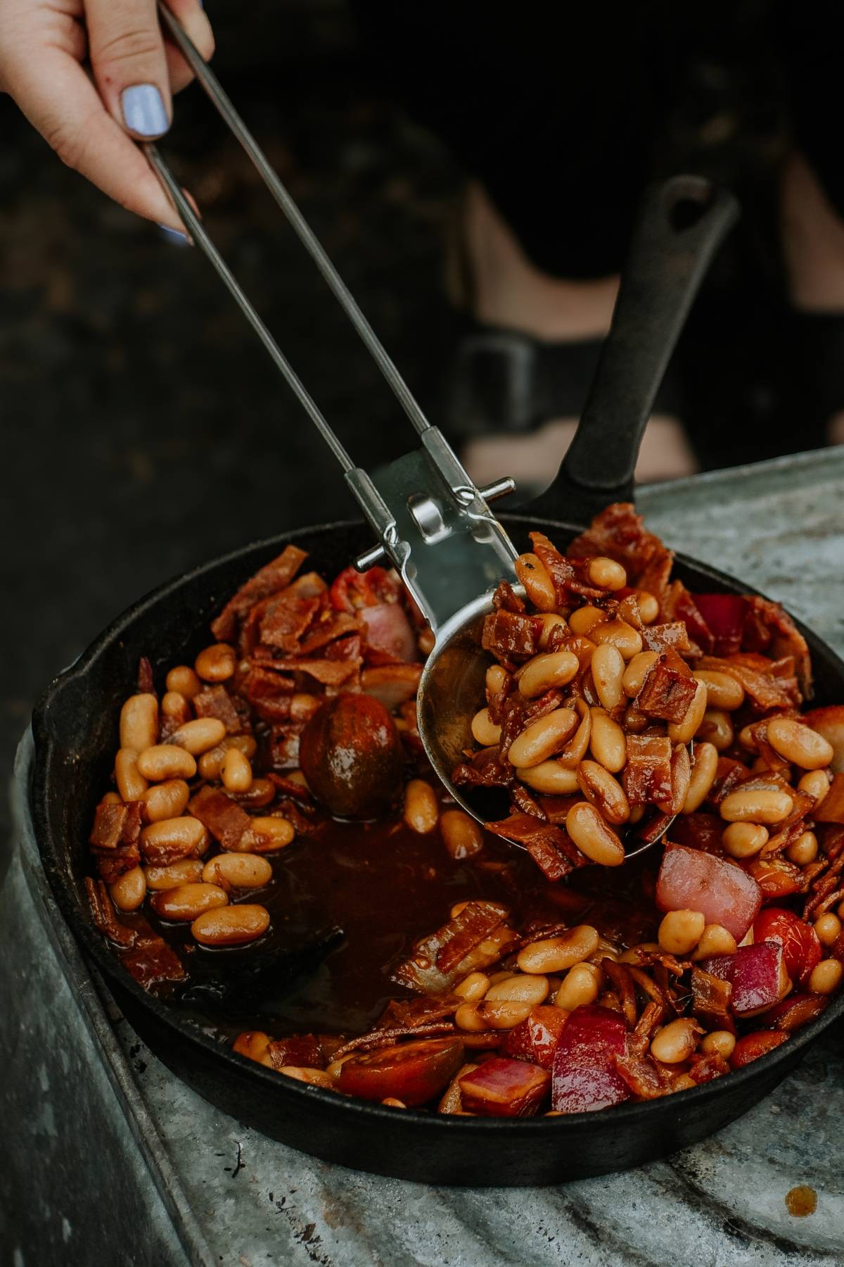 A hand serving baked beans with bacon and onions from a cast iron skillet using metal tongs.