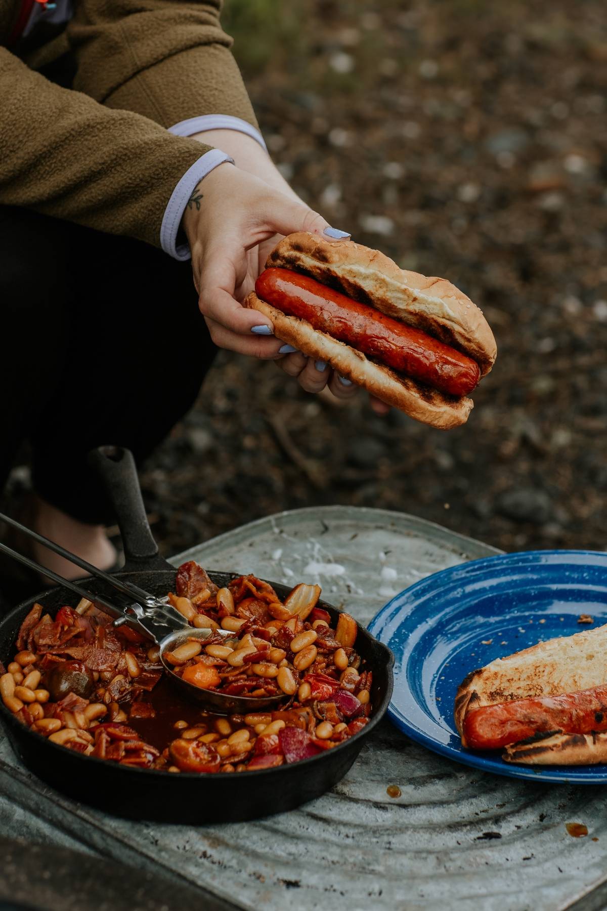 A person holds a sausage hot dog over a skillet of beans and sausage and a blue plate outdoors.