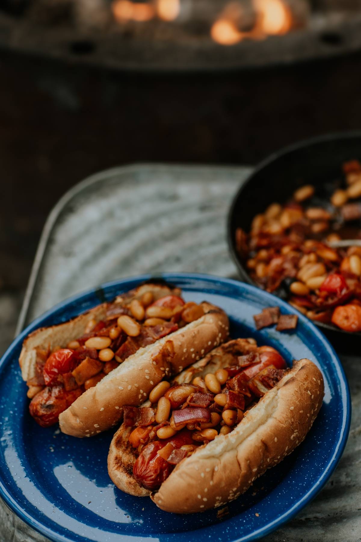 Two hot dogs topped with baked beans and vegetables on a blue plate, with a skillet of toppings nearby.