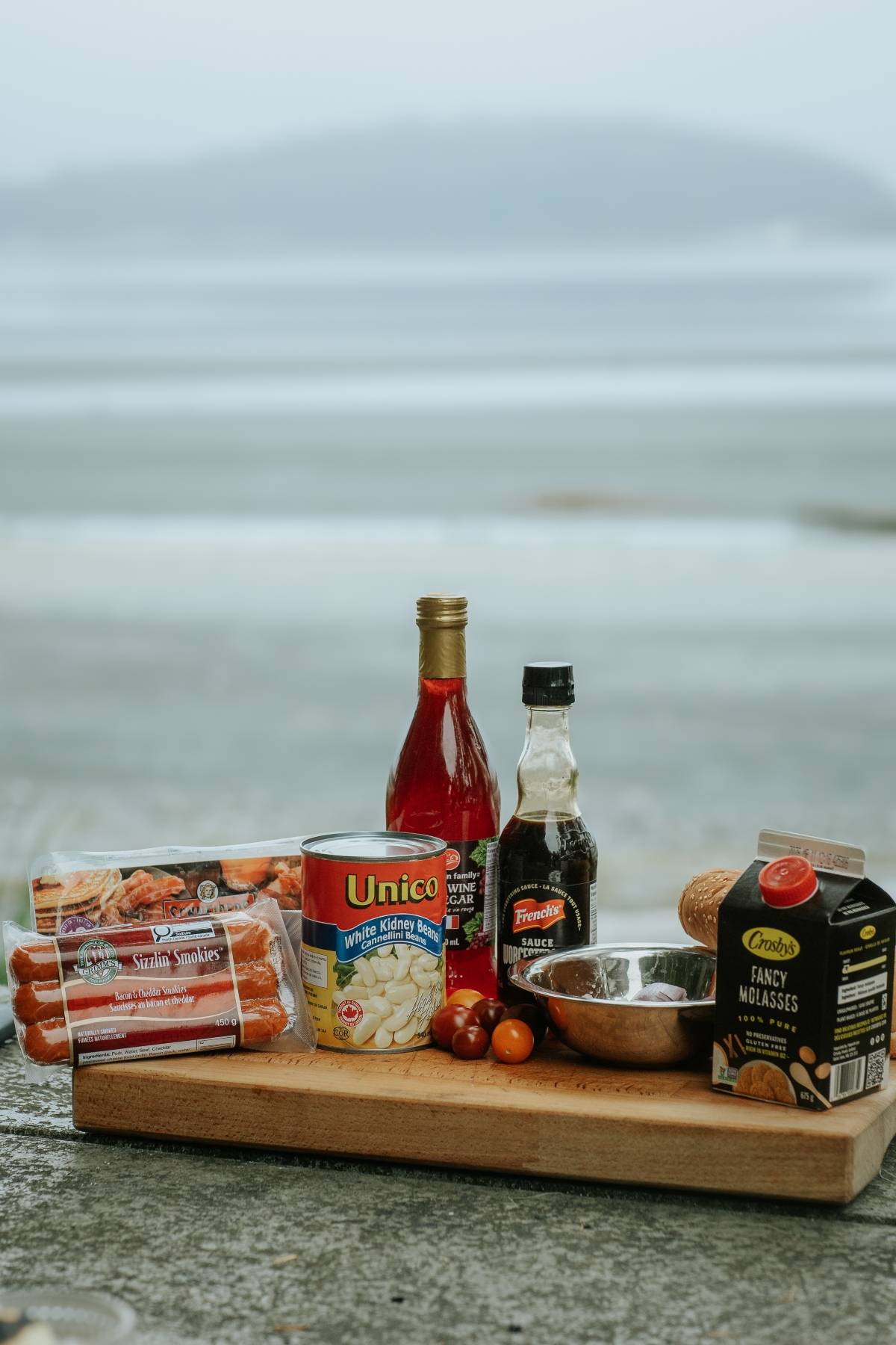 Various cooking ingredients, including beans, sausages, sauces, and tomatoes, arranged on a wooden board outdoors.
