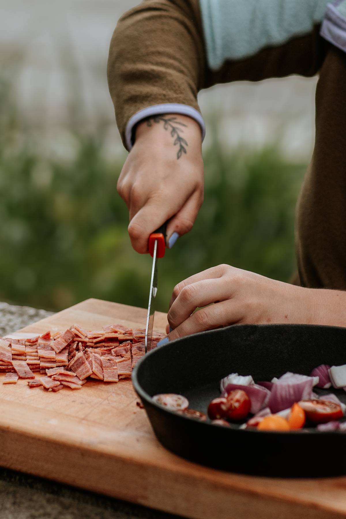 A person slices meat on a wooden board next to a pan with chopped onions and tomatoes.