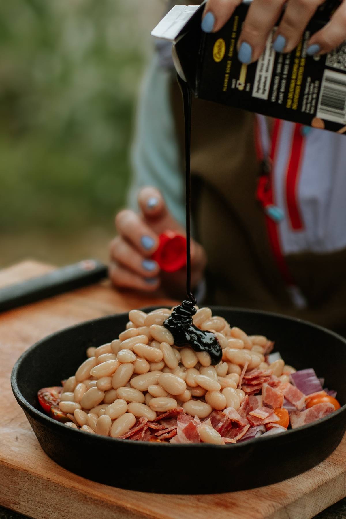 A person pours sauce over white beans, bacon, and vegetables in a black skillet.