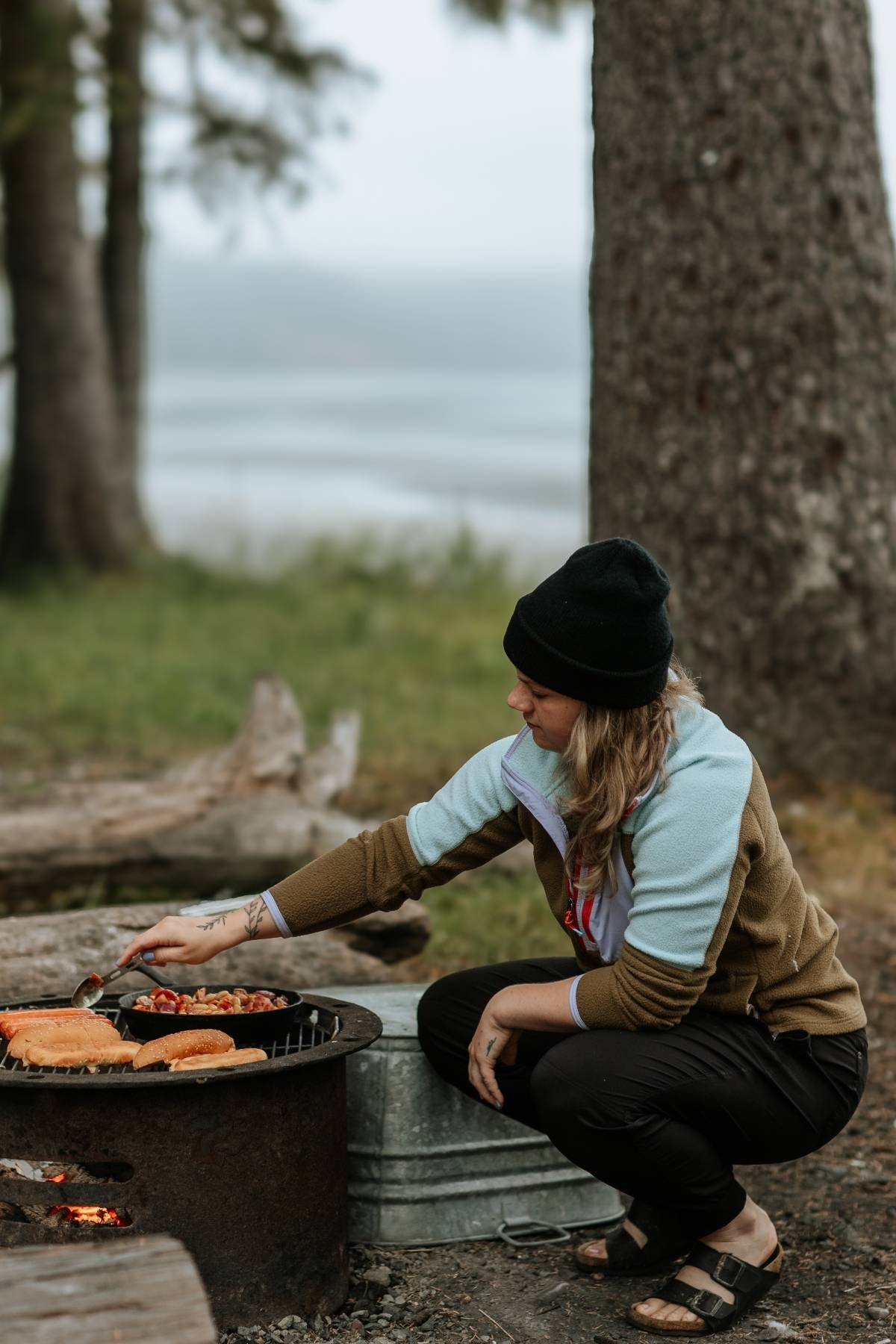 A person in outdoor clothes cooks food on a grill at a campsite near trees and a lake.