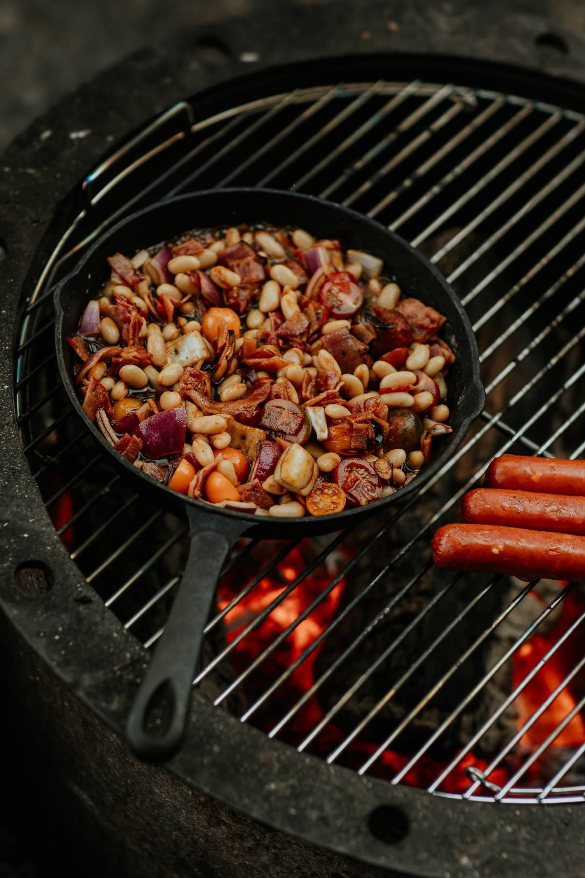 A skillet of beans and vegetables and hot dogs on a grill over an open flame.