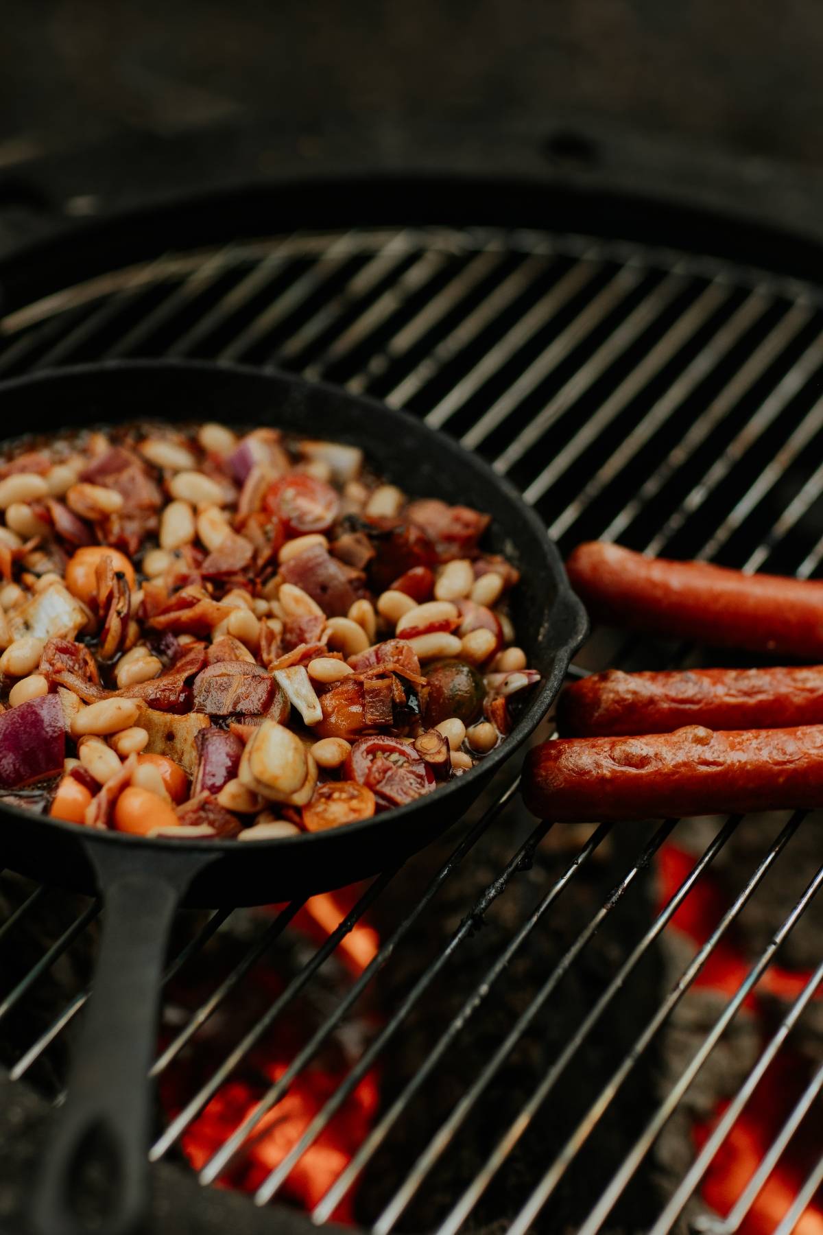 Cast iron skillet with beans and sausages cooking on a grill over an open flame.
