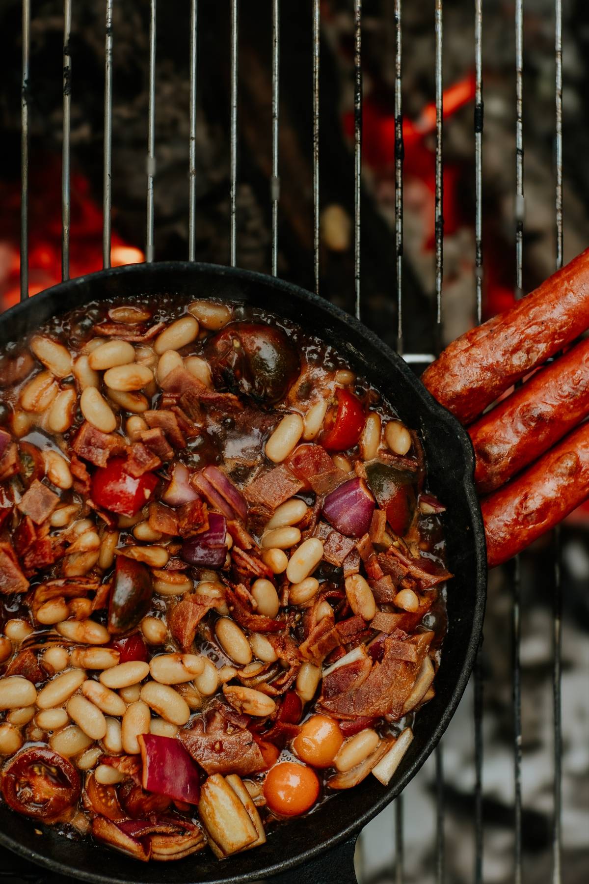 A cast-iron skillet with beans and vegetables cooking on a grill beside grilled sausages.