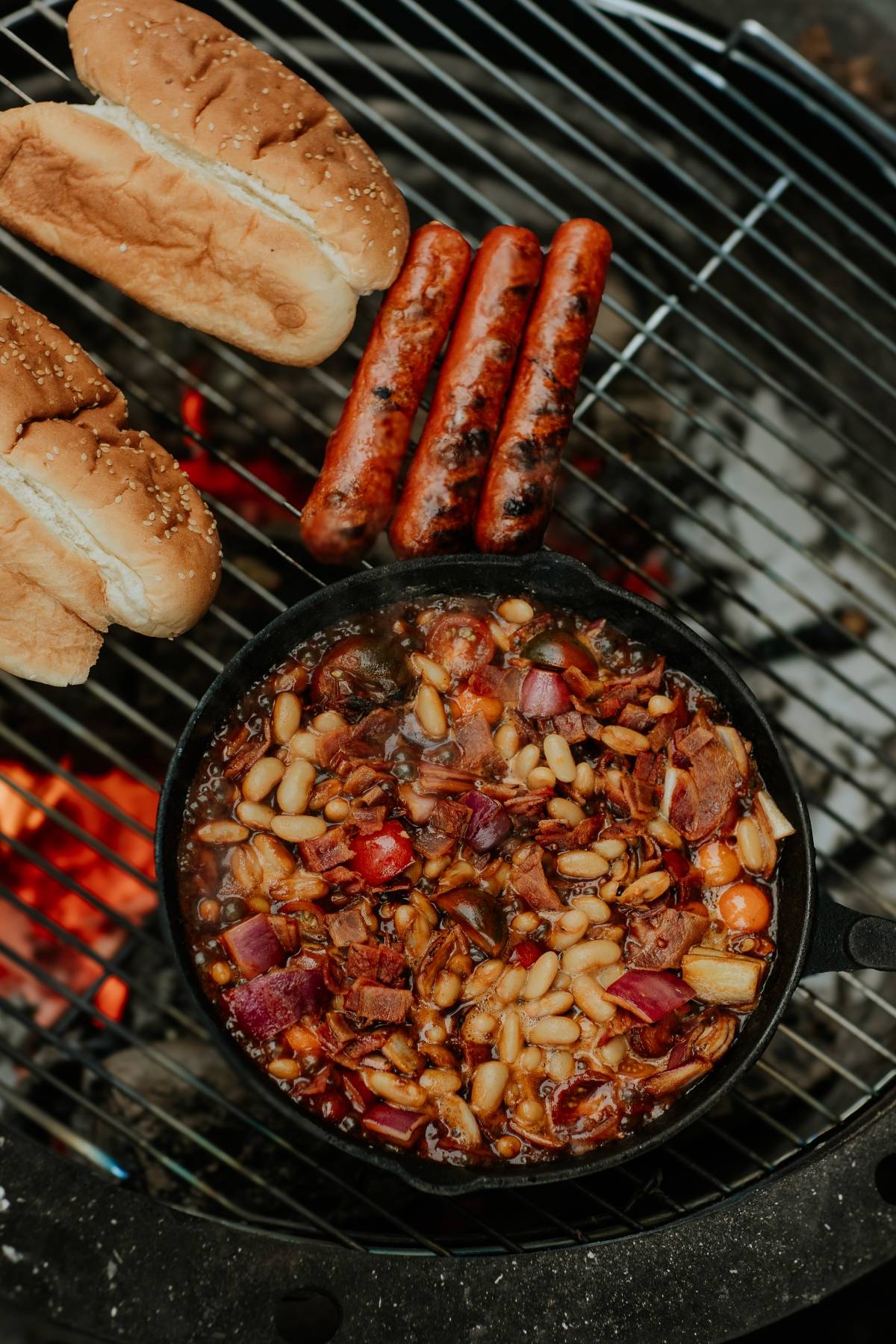 Hot dogs and buns on a grill next to a cast iron skillet of baked beans over an open flame.