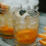 Two glasses with campfire marshmallow cocktails, ice, toasted marshmallows, and orange slices beside them on a gray surface.