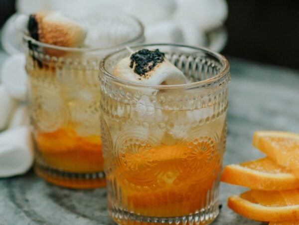 Two glasses with campfire marshmallow cocktails, ice, toasted marshmallows, and orange slices beside them on a gray surface.