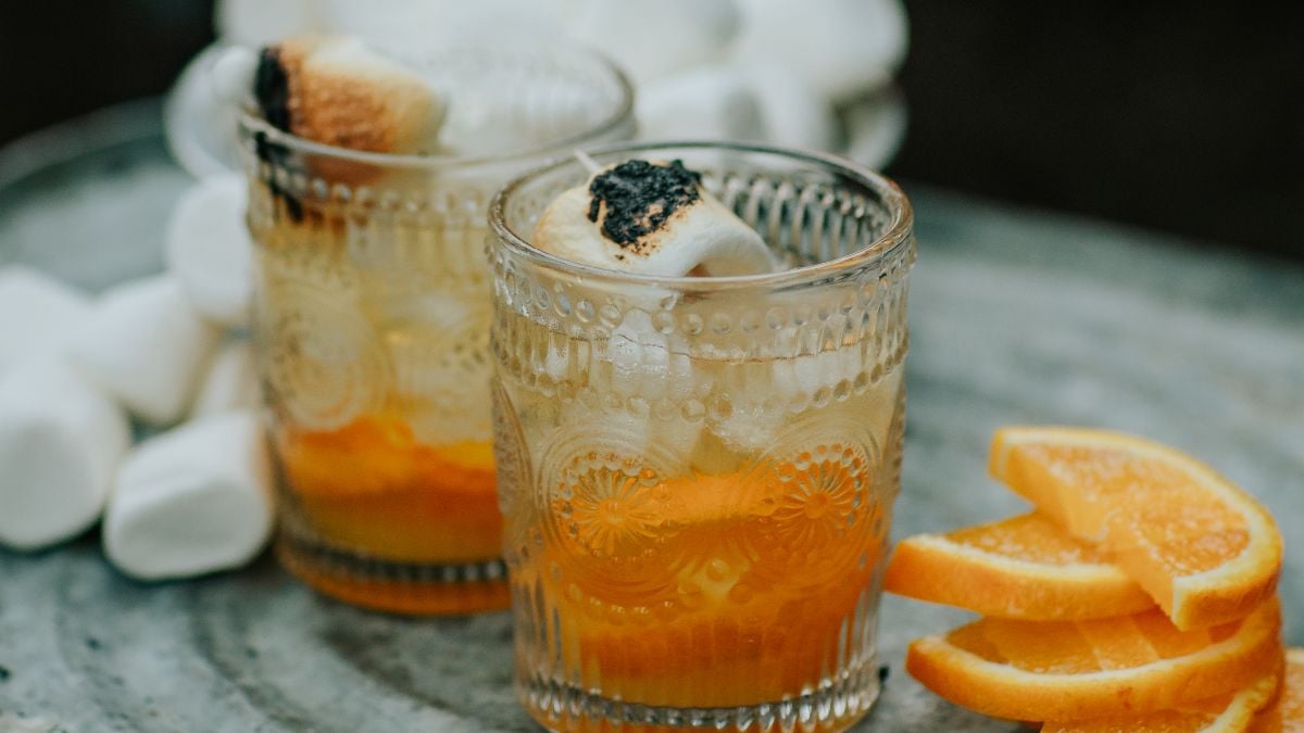 Two glasses with campfire marshmallow cocktails, ice, toasted marshmallows, and orange slices beside them on a gray surface.