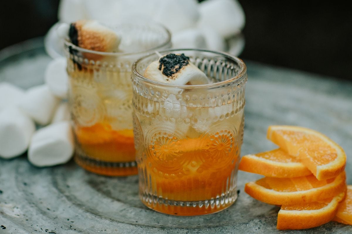 Two glasses of iced orange campfire marshmallow cocktail, garnished with toasted marshmallows, sit beside fresh orange slices and fluffy marshmallows.