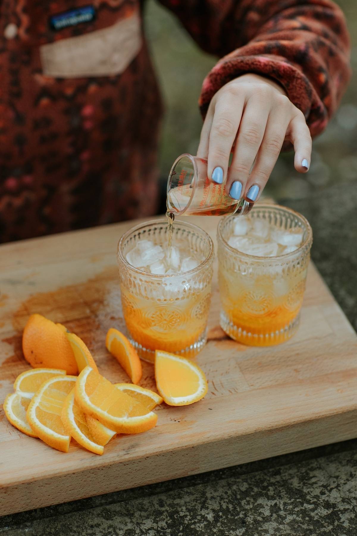 A person pours liquid into a glass with ice and orange slices on a wooden board with more orange slices.