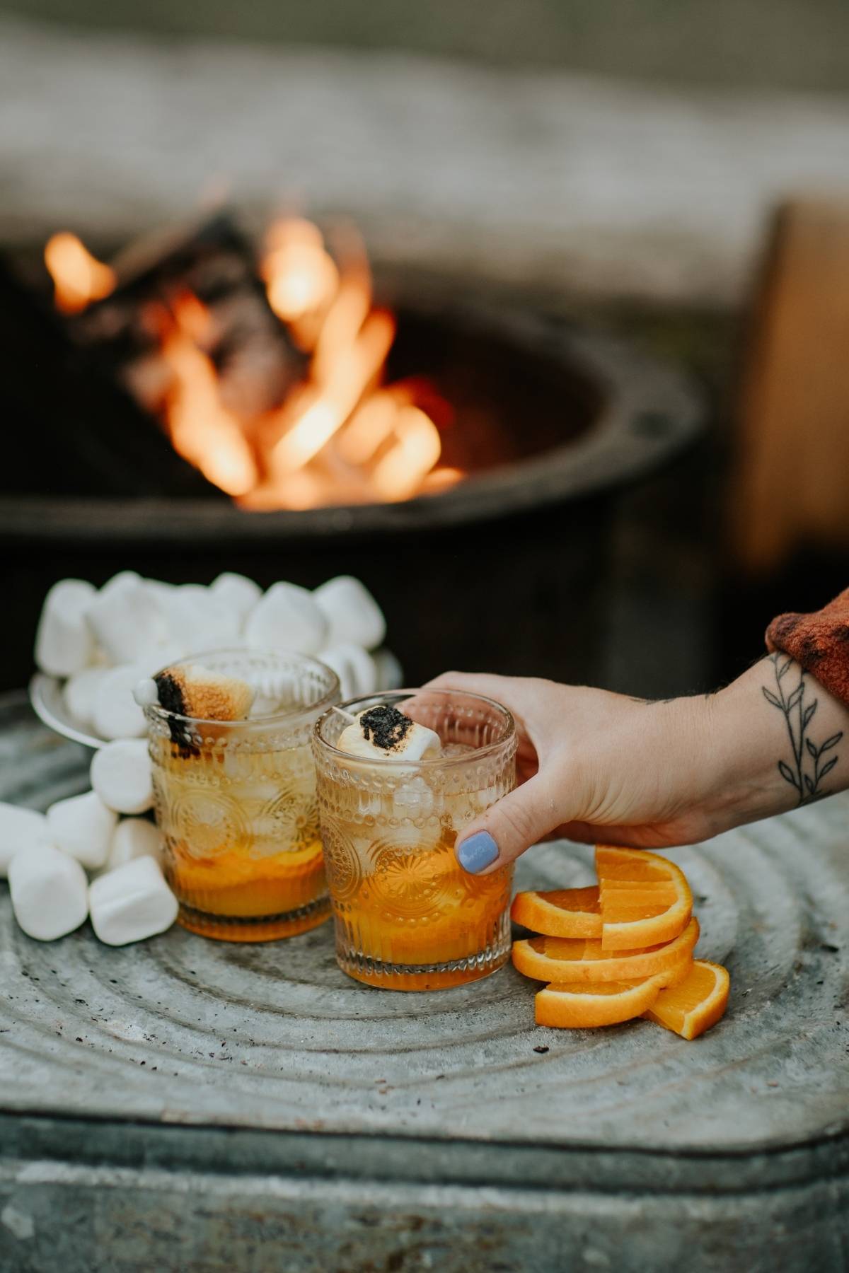 Two glasses with orange drinks and toasted marshmallows, orange slices, and a firepit in the background.