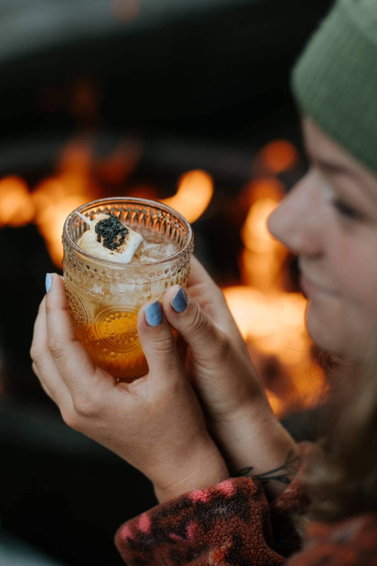 Person holding a drink topped with a toasted marshmallow by a fire, with blurred face and warm background.