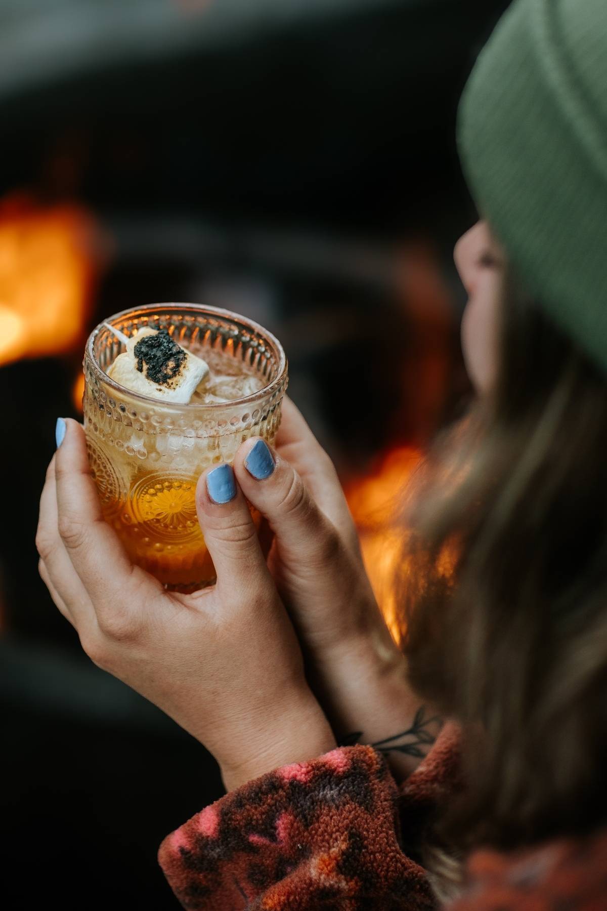 Person holding a decorative glass of drink with a toasted marshmallow and topping, near a fire.