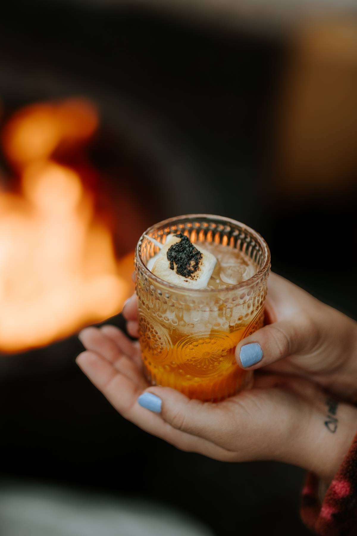 A hand holds a cocktail with ice, garnished with a toasted marshmallow and black topping.