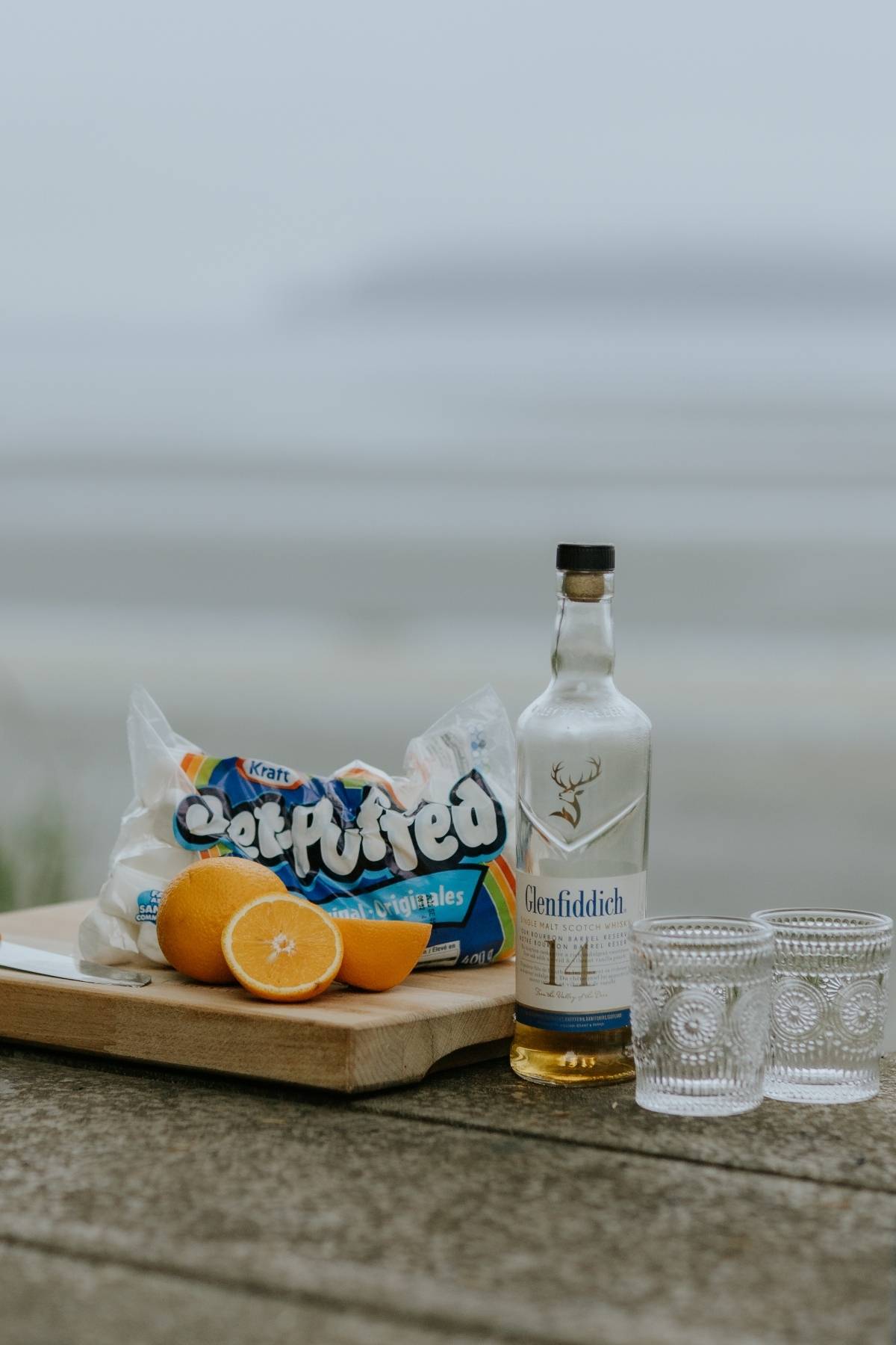 Bottle of Glenfiddich, two glasses, oranges, and marshmallows on a board by the beach.