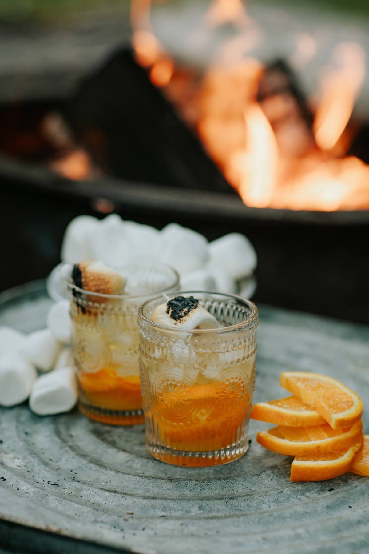 Two glasses with toasted marshmallows, orange slices, and ice, set by a campfire with marshmallows nearby.
